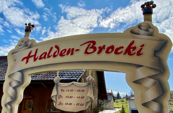 Die Tafel von der Halden-Brocki mit dem Logo und den Öffnungszeiten steht vor dem Haus. Sie stammt vom Künstler Andy Ineichen – Illusions- und Kunstmalerei, Steinhausen.